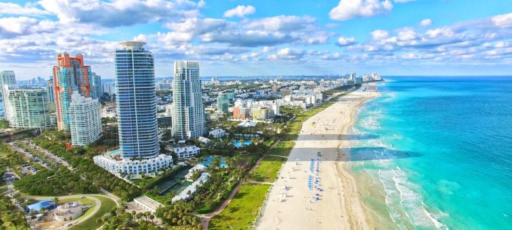 This image shows a stunning aerial view of Sunny Isles Beach, Florida, characterized by its pristine white sandy beaches and vibrant turquoise waters stretching along the coastline. The picture highlights luxury high-rise buildings with modern architectural designs, surrounded by lush green landscaping and recreational areas. The blue skies with scattered fluffy clouds add to the tropical charm, making it a picturesque representation of the upscale and serene lifestyle that Sunny Isles Beach is known for.
